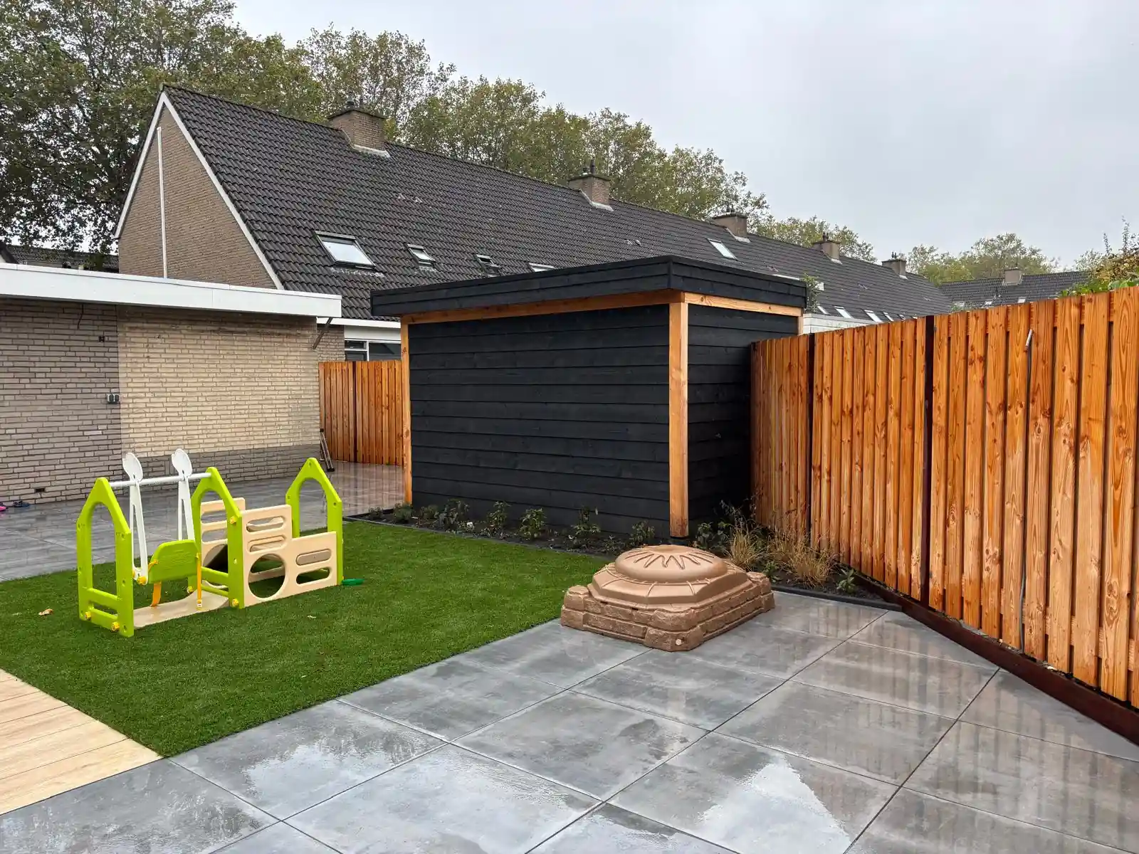 Zwarte houten schuur met plat dak naast een veld kunstgras met een groen speeltoestel, met een terras van grote grijze tegels en een zandbak in schelpvorm