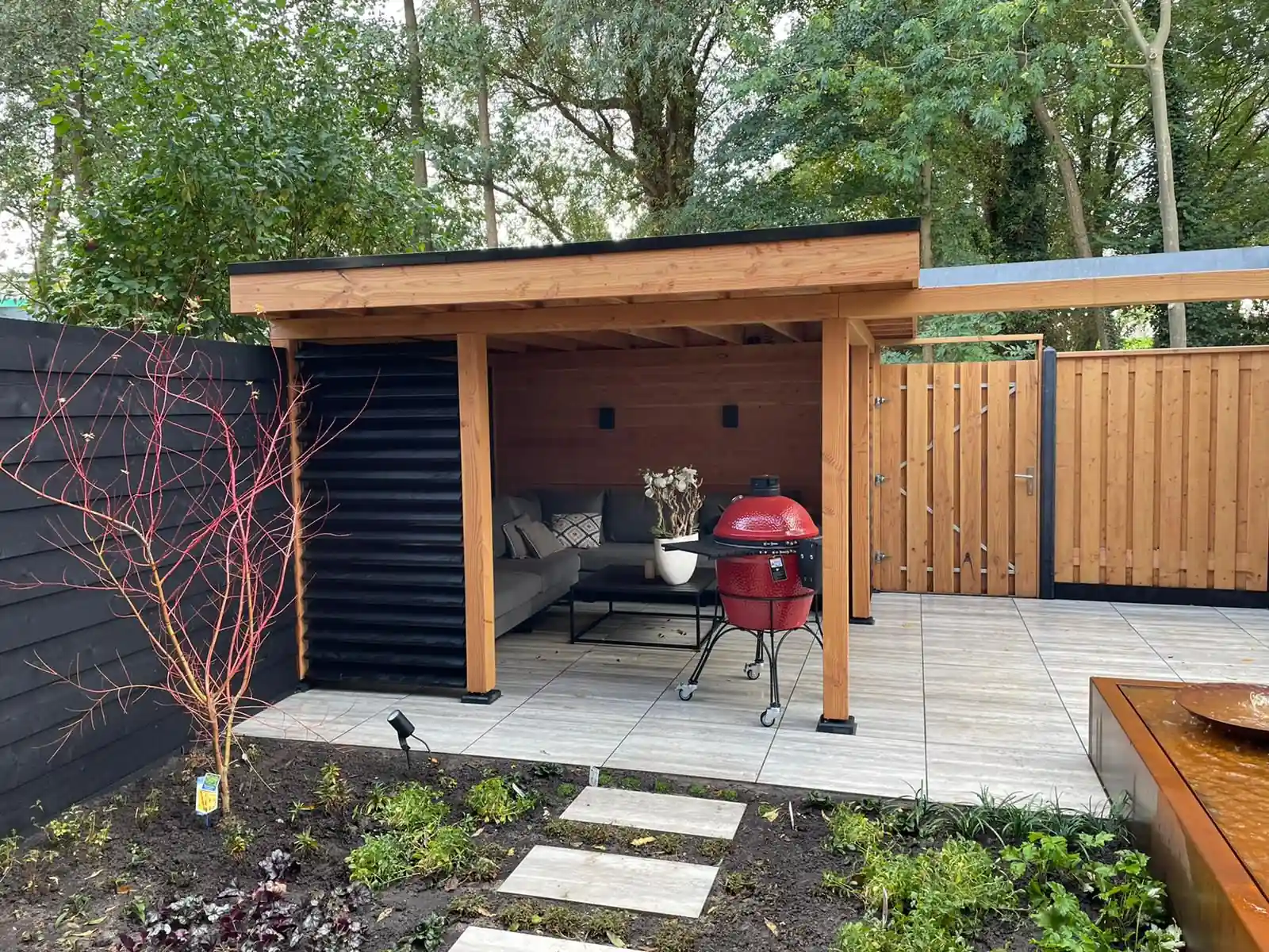 Douglas overkapping met zwarte lamellenwand en loungebank naast een houten poort, met een border met jonge beplanting en stapstenen ervoor