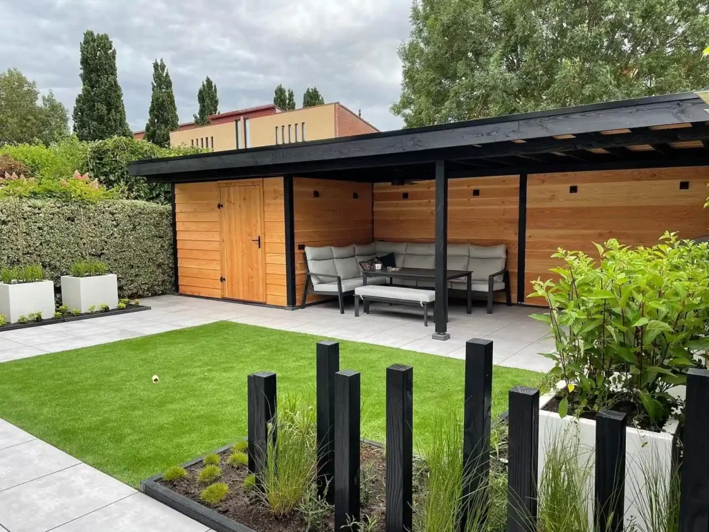Overzicht van de achtertuin met een Douglas overkapping met loungeset en schuur, een veld kunstgras en borders met witte plantenbakken en zwarte palen
