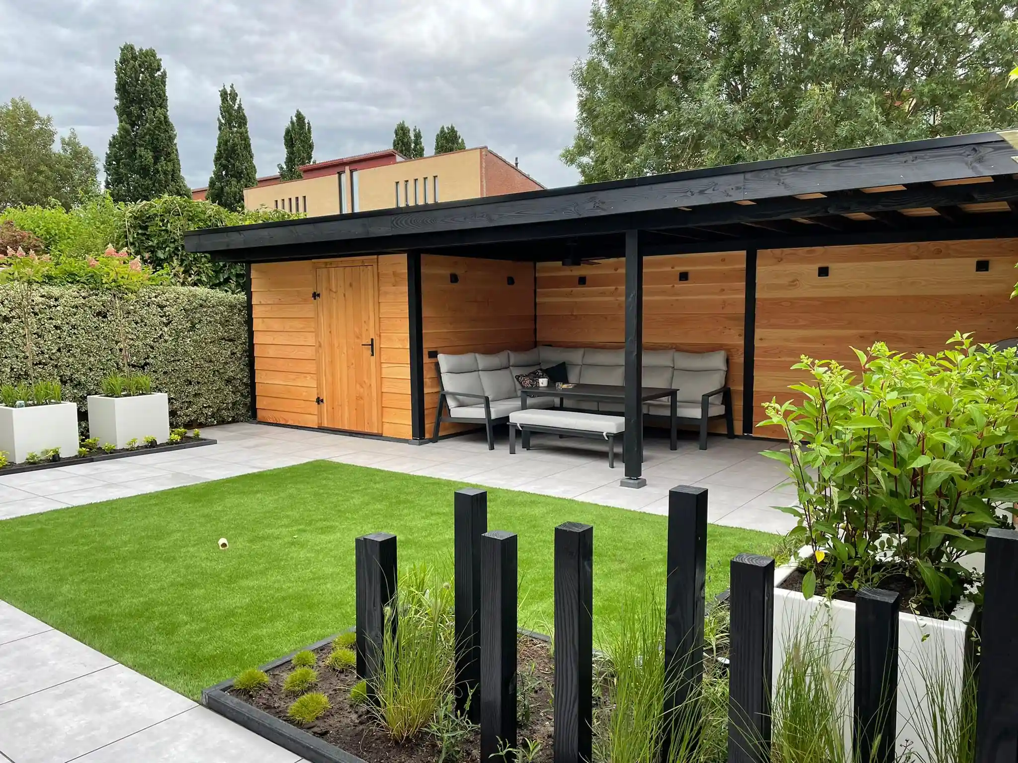 Douglas overkapping met zwarte staanders en aangebouwde schuur met houten deur, met een loungeset, een veld kunstgras en een border met zwarte palen en siergrassen