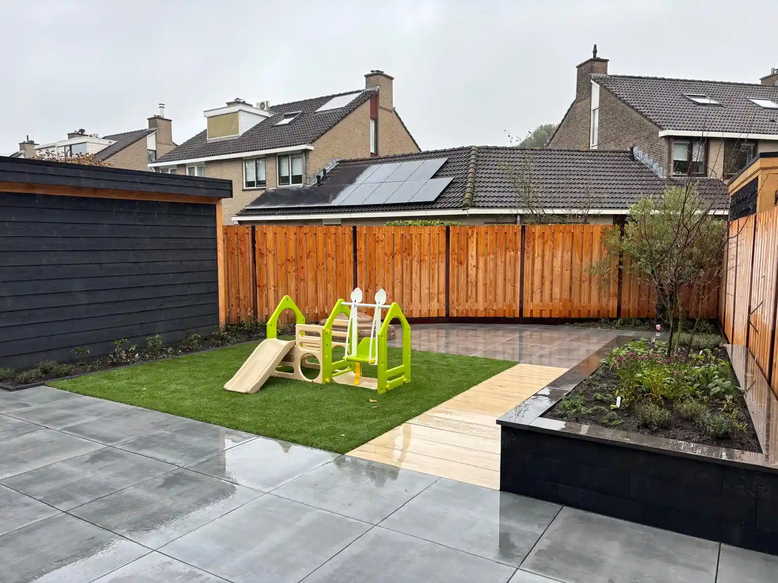 Overzicht van de achtertuin met een nieuwe houten schutting rond een speelhoek van kunstgras, met een zwarte schuurwand, tegelterras en verhoogde border