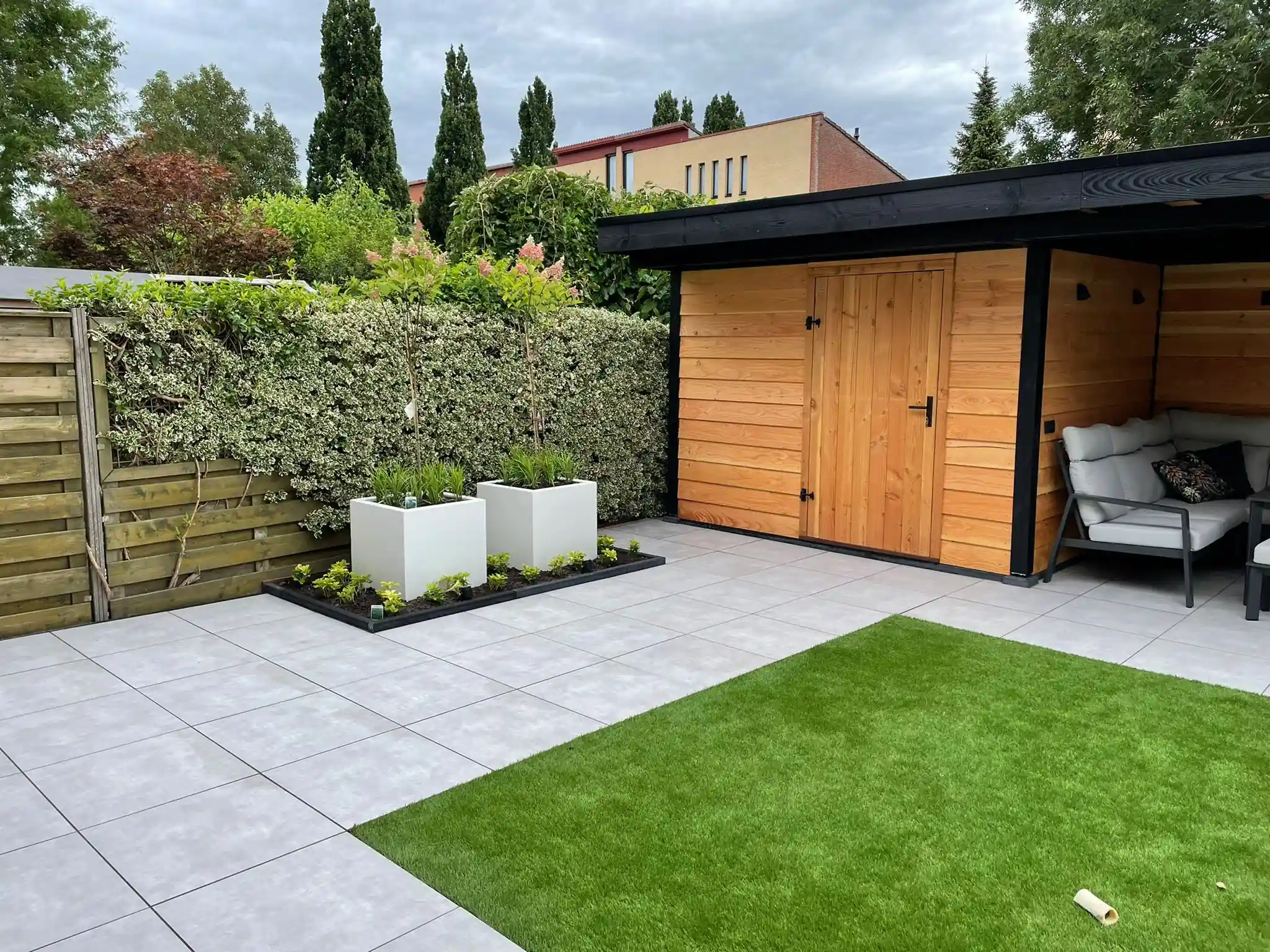 Schuur met Douglas rabatdelen en een houten deur onder een zwarte overkapping met loungeset, met een terras van lichtgrijze tegels en een veld kunstgras