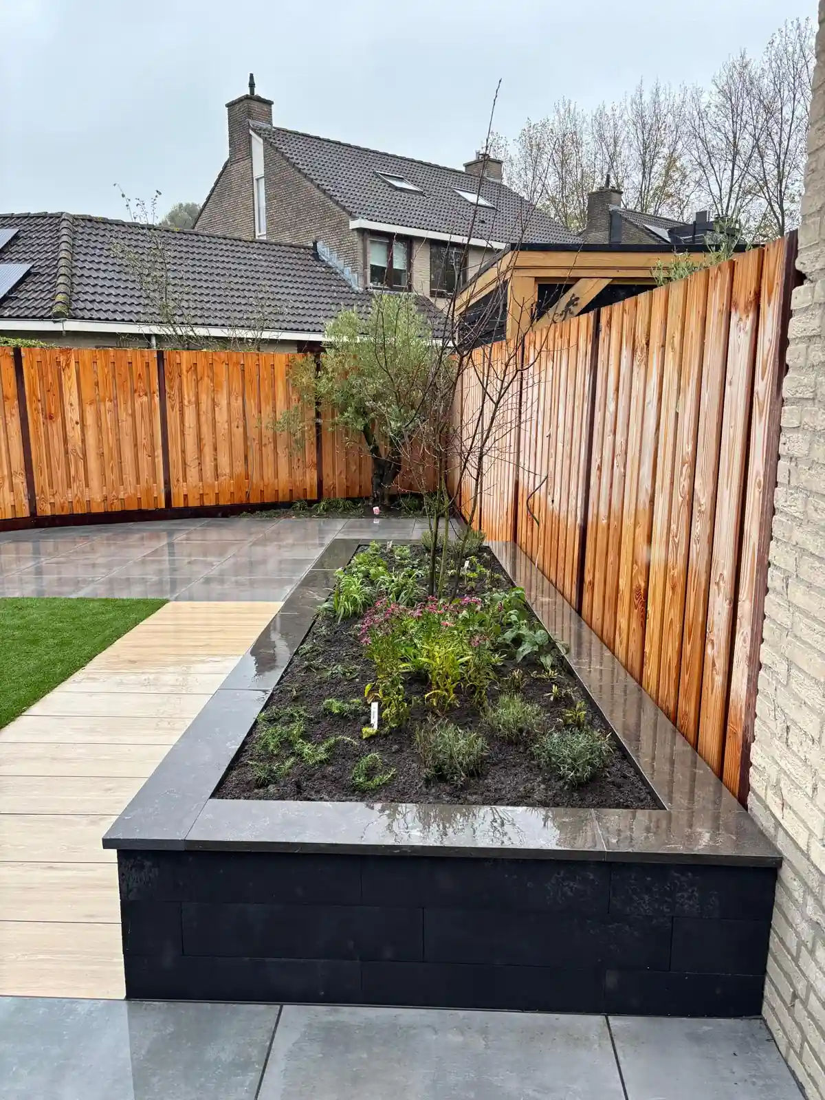 Verhoogde border van zwarte stapelblokken met een gepolijste afdekrand, gevuld met verse beplanting en een jonge boom, langs een pad van houtlook tegels