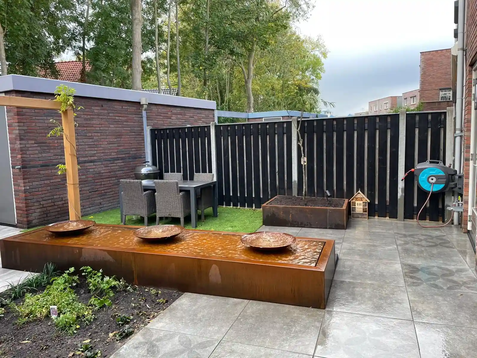 Cortenstalen waterelement met drie schalen langs een terras, met daarachter een eethoek met rieten stoelen op kunstgras en een zwarte schutting met plantenbak