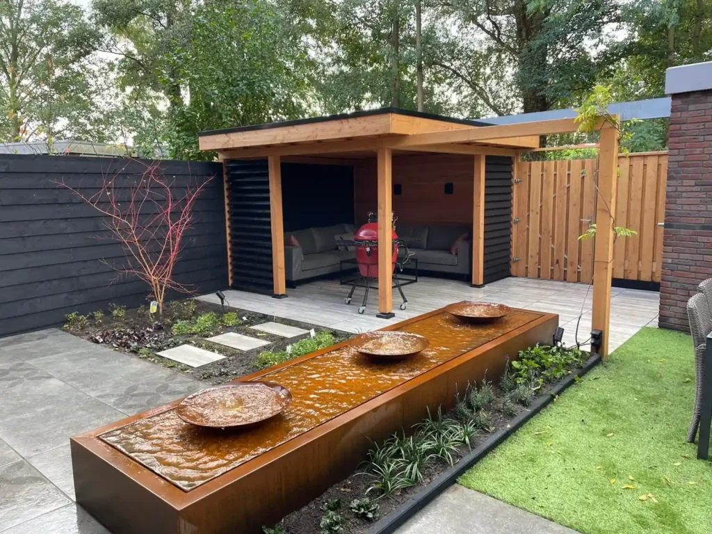 Overzicht van de achtertuin met een cortenstalen waterbak met drie schalen, een Douglas overkapping met loungebank en borders met verse beplanting