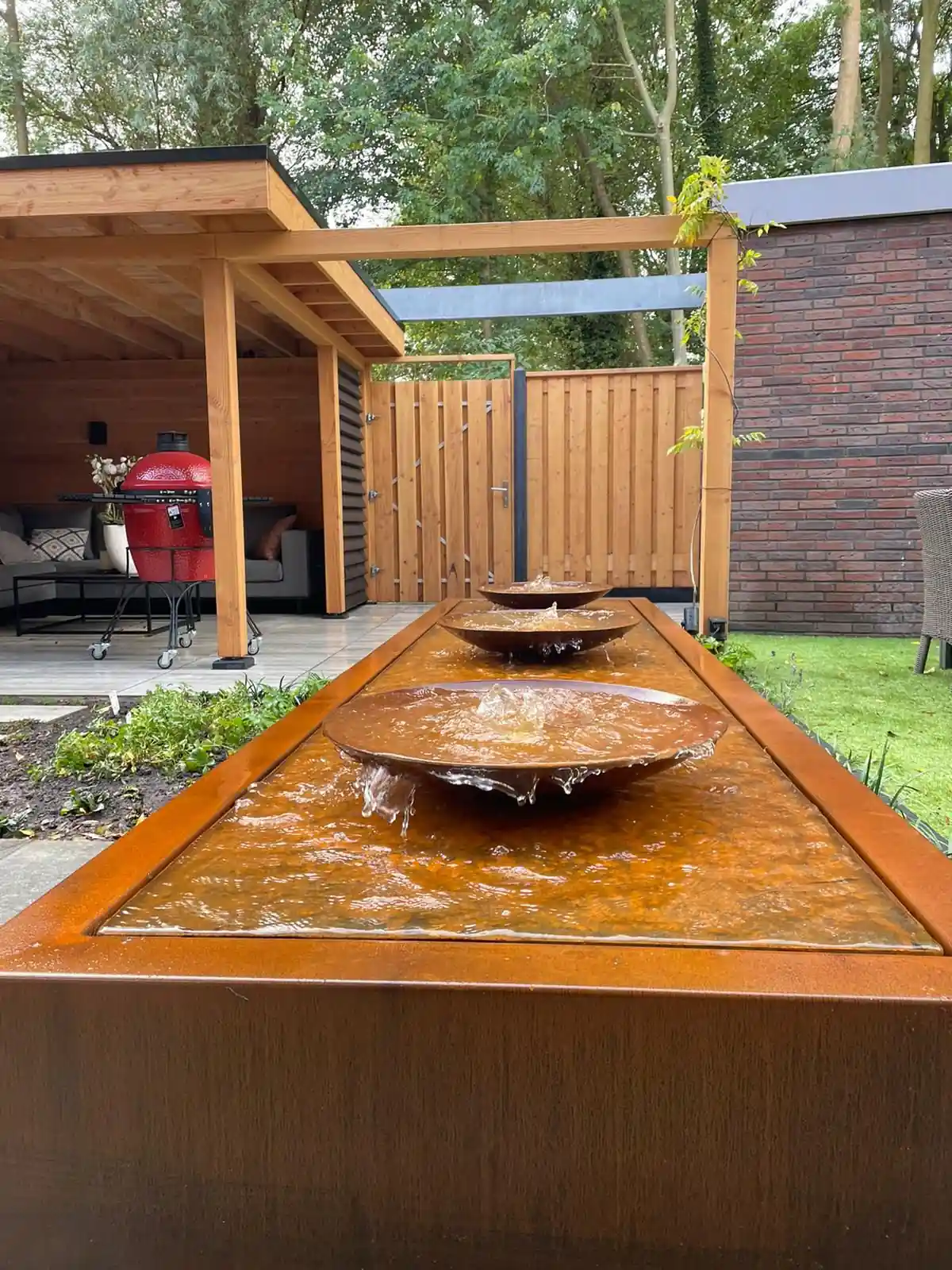 Drie cortenstalen waterschalen die overlopen in een langgerekte waterbak, met de Douglas overkapping en de rode barbecue op de achtergrond