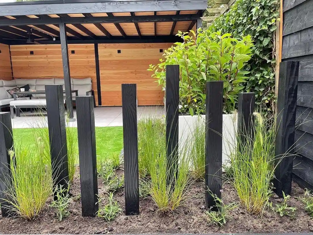 Rij zwart gebrande houten palen op verschillende hoogtes tussen verse siergrassen in een border, met de overkapping en het kunstgras op de achtergrond