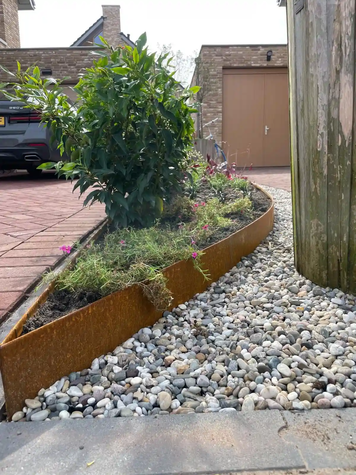 Gebogen cortenstalen rand die een border met laurier en roze bloeiende vaste planten scheidt van een strook ronde keien langs de klinkerbestrating