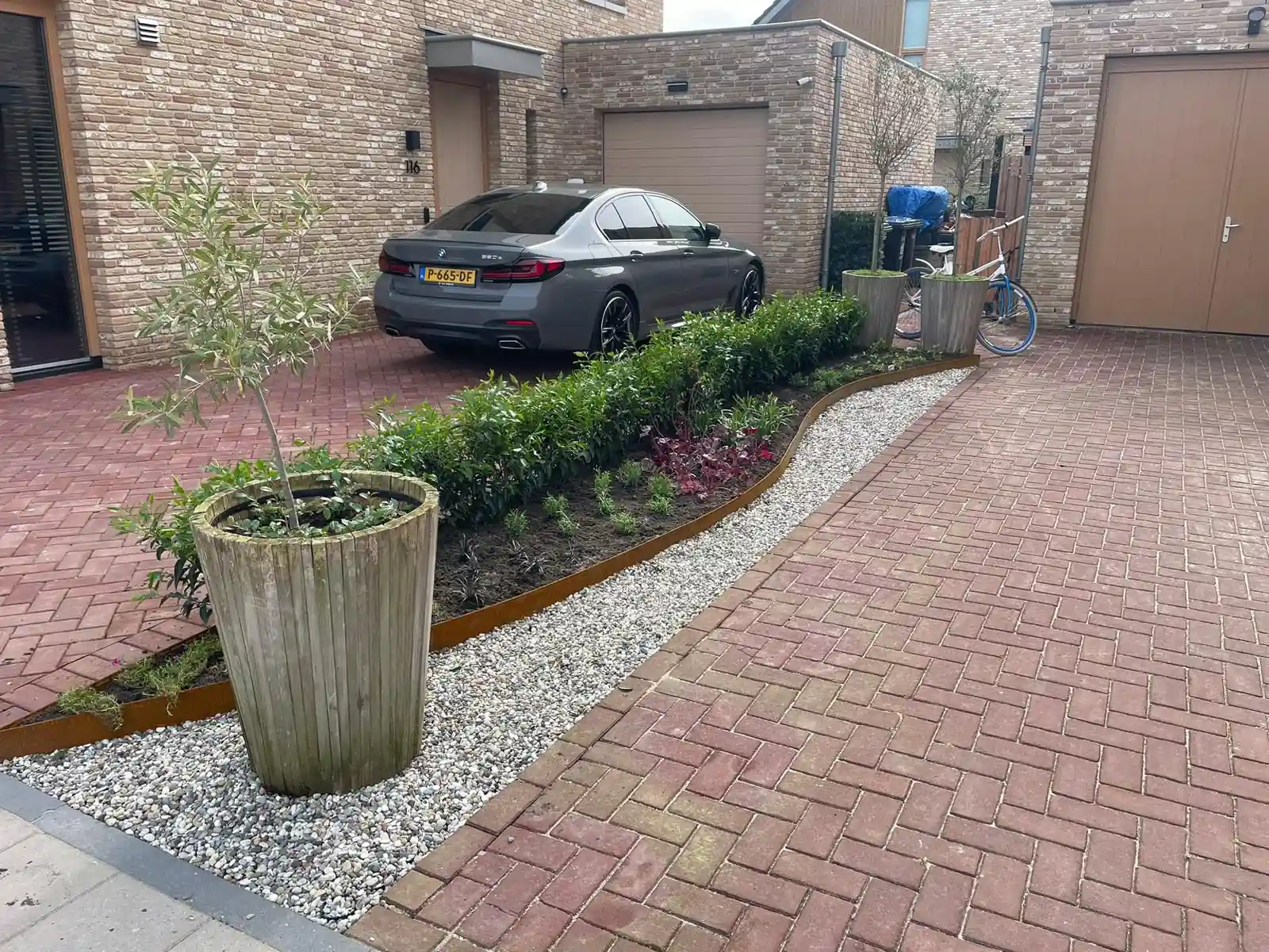 Ruime oprit van roodbruine klinkers in keperverband met een gebogen border achter een cortenstalen rand, een strook keien en een olijfboom in een houten pot