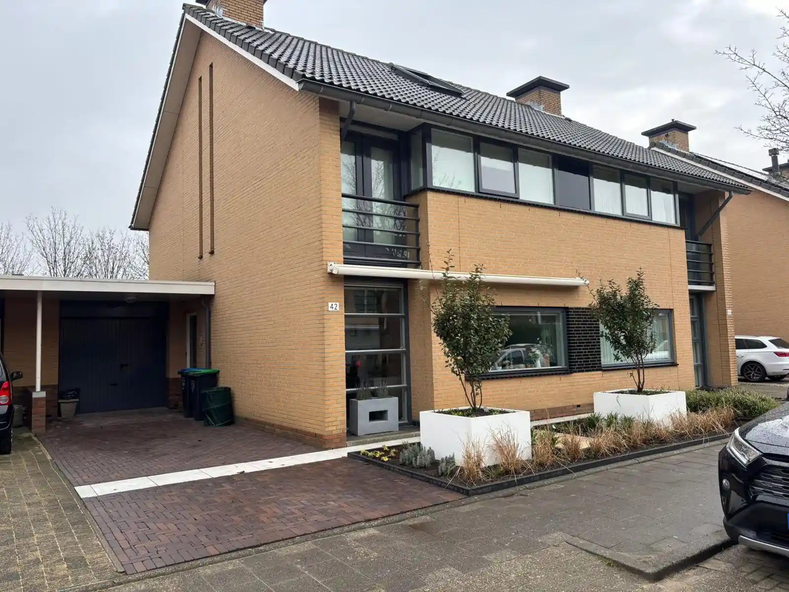 Vooraanzicht van de woning met een bestrate oprit van donkere klinkers naar de carport, met een verse border en witte plantenbakken langs de gevel