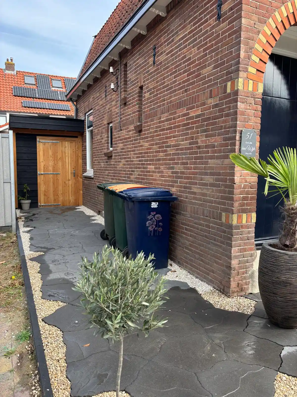Pad van antracietkleurige flagstones met een strook wit grind langs een bakstenen gevel, met afvalcontainers, een houten bergingsdeur en een jong olijfboompje