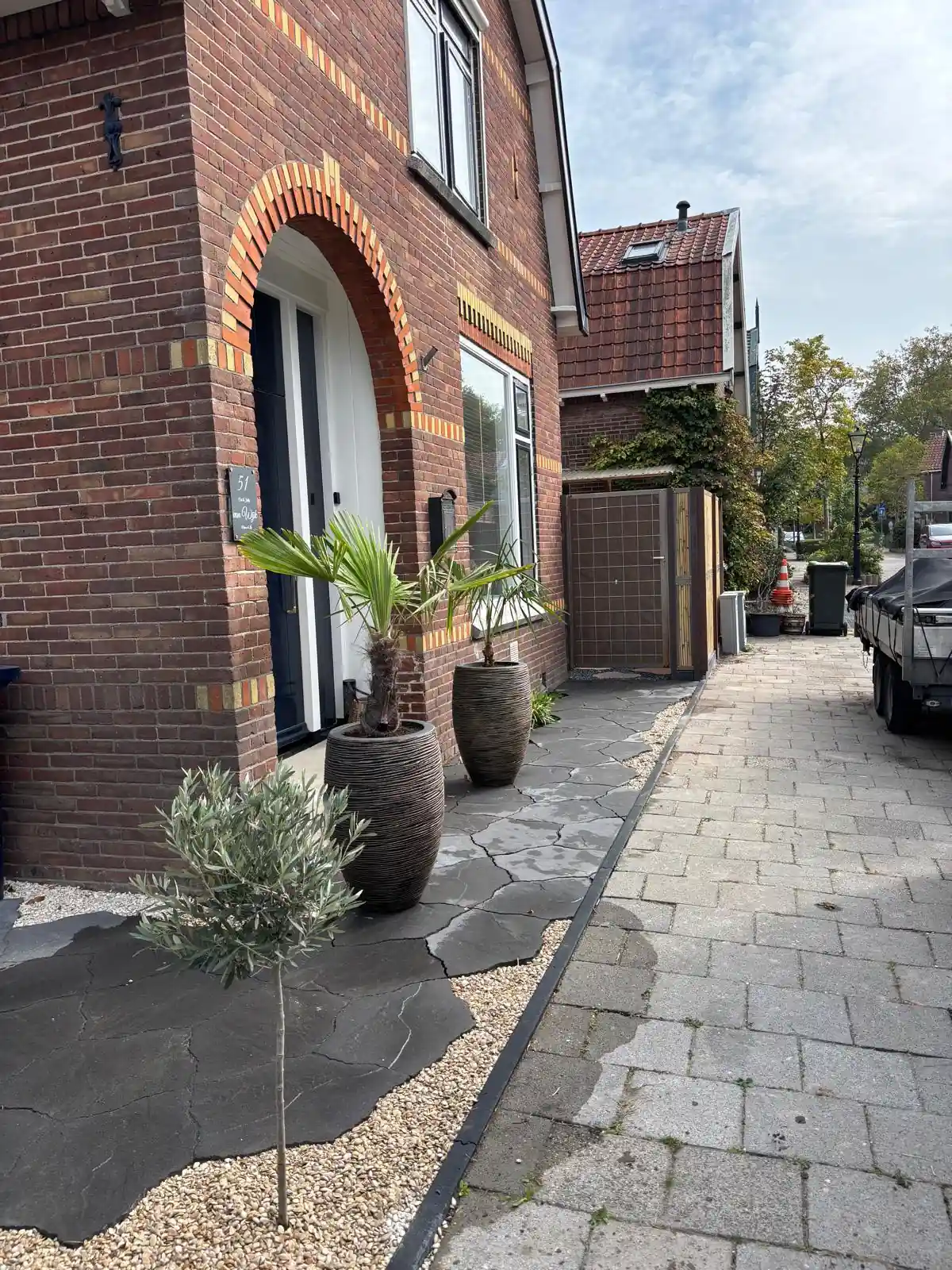Pad van antracietkleurige flagstones langs de voorgevel met een boogvormige entree, met twee palmen in ribbelpotten, een olijfboompje en een strook grind