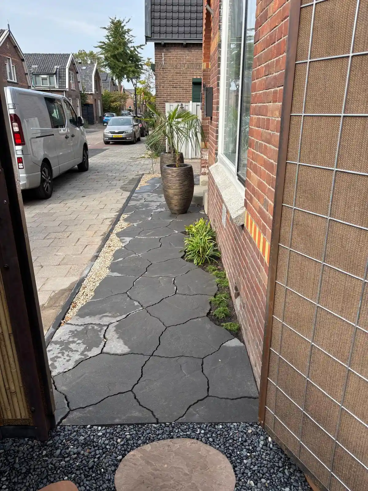 Pad van antracietkleurige flagstones langs de voorgevel richting de straat, met een strook grind, beplanting tegen de muur en palmen in ribbelpotten