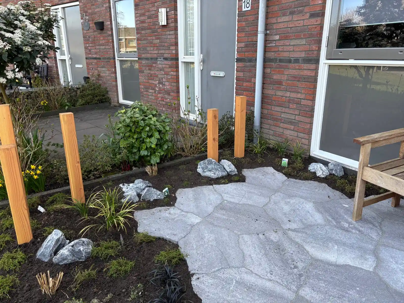 Voortuin met verse beplanting, siergrassen en grijze keien in donkere aarde, rondom een terras van flagstones met Douglas palen