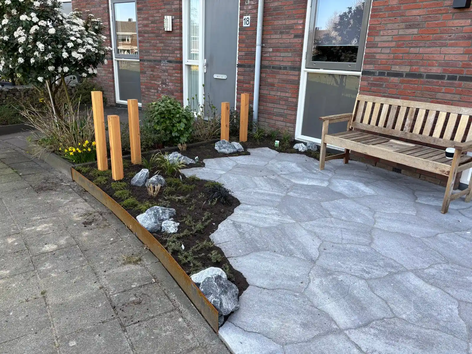 Border met bloeiende narcissen, kruipplanten en grijze keien langs een terras van flagstones, met een rij Douglas palen en een houten bank