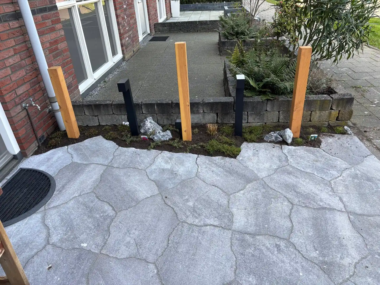 Terras van grijze flagstones langs een lage keermuur, met Douglas palen, zwarte staande tuinlampen en een smalle border met keien en varens
