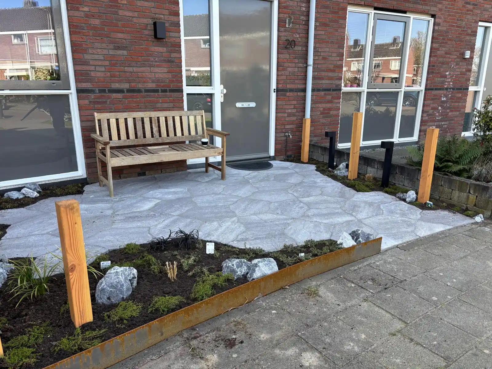 Voortuin met een terras van grijze flagstones, een houten bank bij de voordeur, Douglas palen en een border met keien achter een cortenstalen rand
