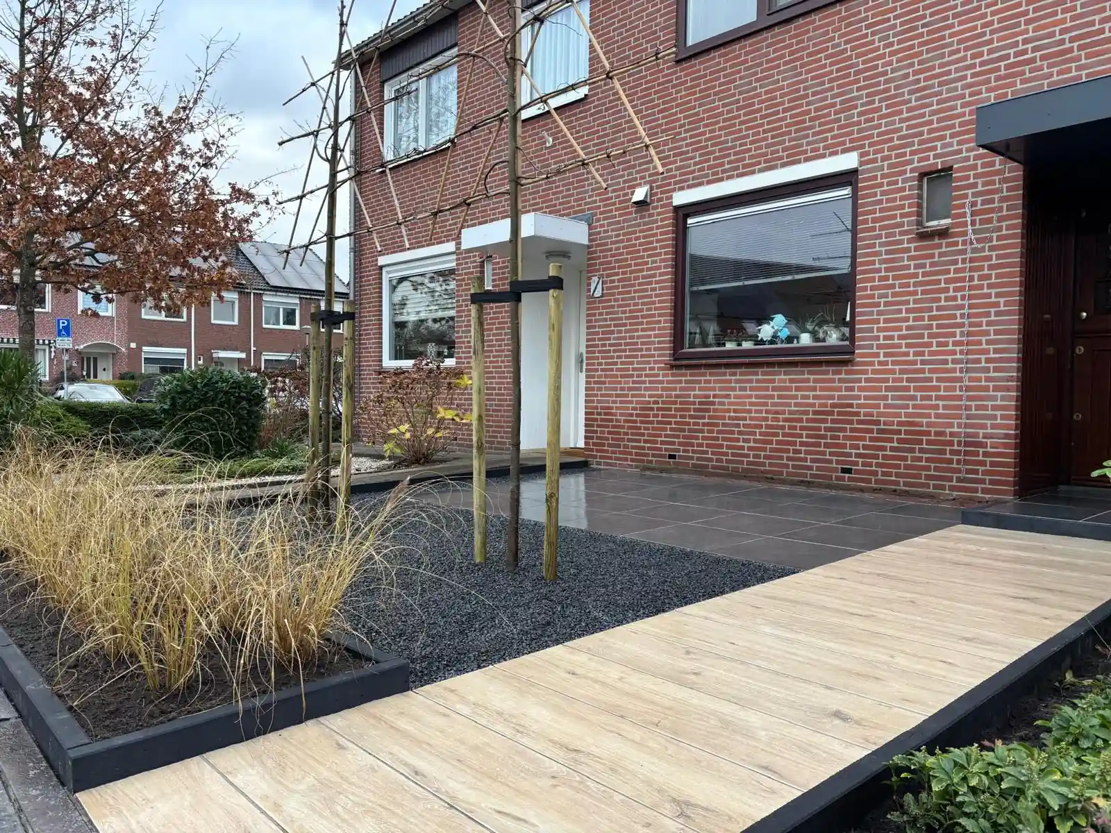 Pad van lichte houtlook tegels langs een border met siergrassen achter een antracieten kantopsluiting, met leibomen op palen in een vak zwart split