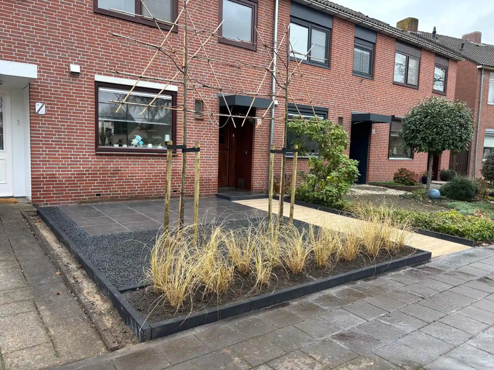 Overzicht van de voortuin met een terras van grijze tegels, een vak zwart split met leibomen en een brede border met siergrassen langs het trottoir
