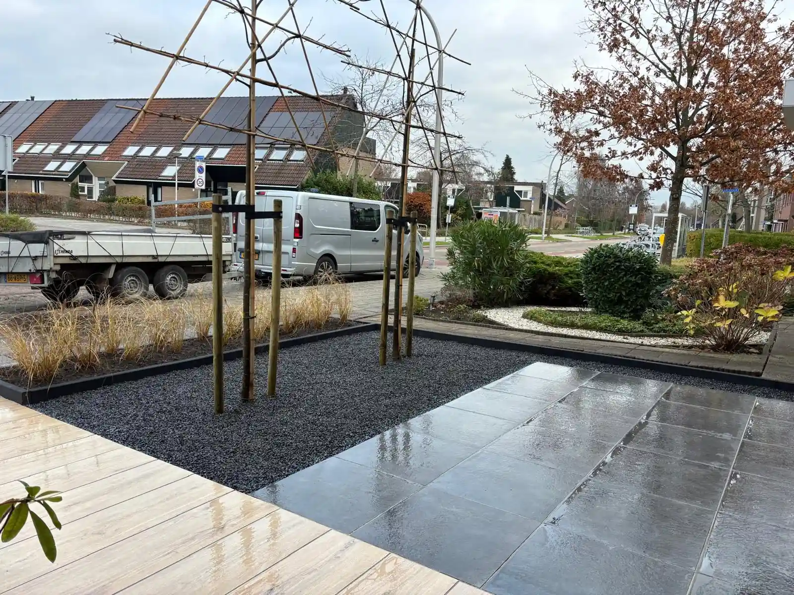 Vak zwart split met leibomen op boompalen, afgezet met een antracieten rand, tussen een terras van grijze tegels en een pad van lichte houtlook tegels