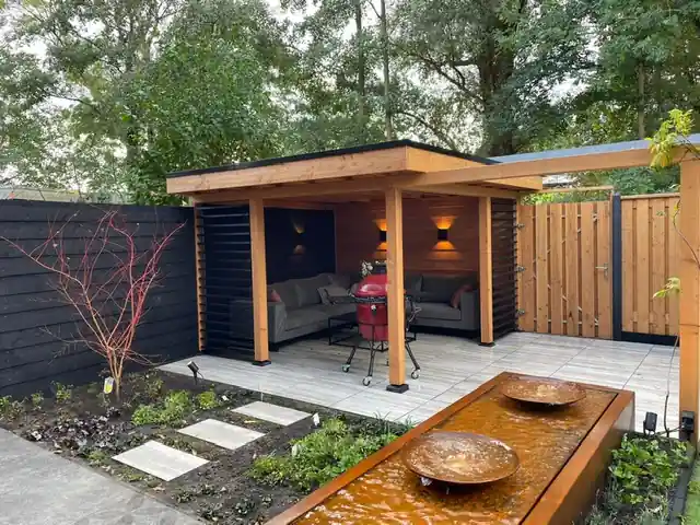 Verlichte Douglas overkapping met loungebank en rode keramische barbecue, met daarvoor een cortenstalen waterbak met twee schalen en stapstenen
