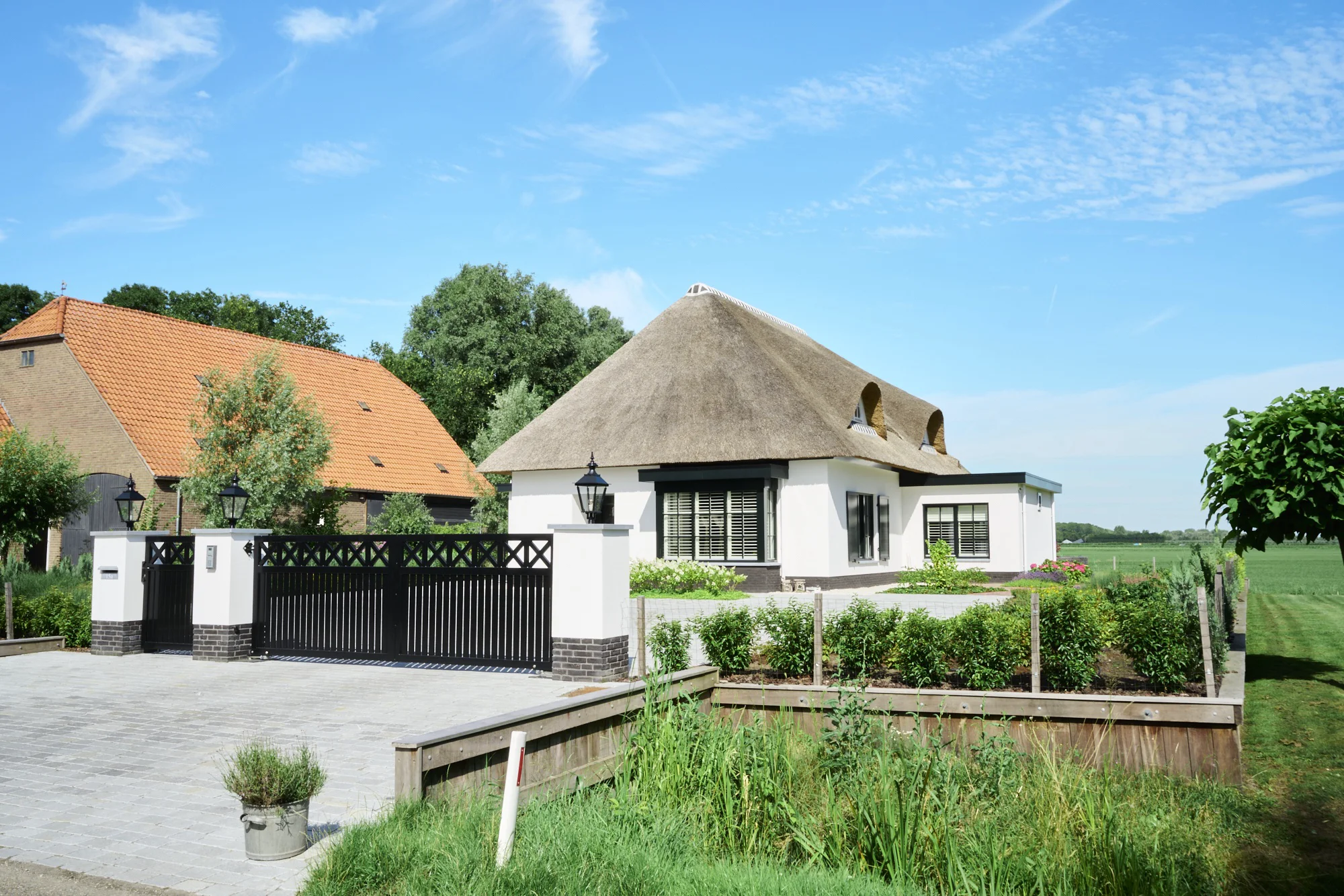 Ruime bestrate oprit met een zwart toegangshek tussen witte pilaren, jonge haagbeplanting in houten bakken en een woning met rieten kap erachter