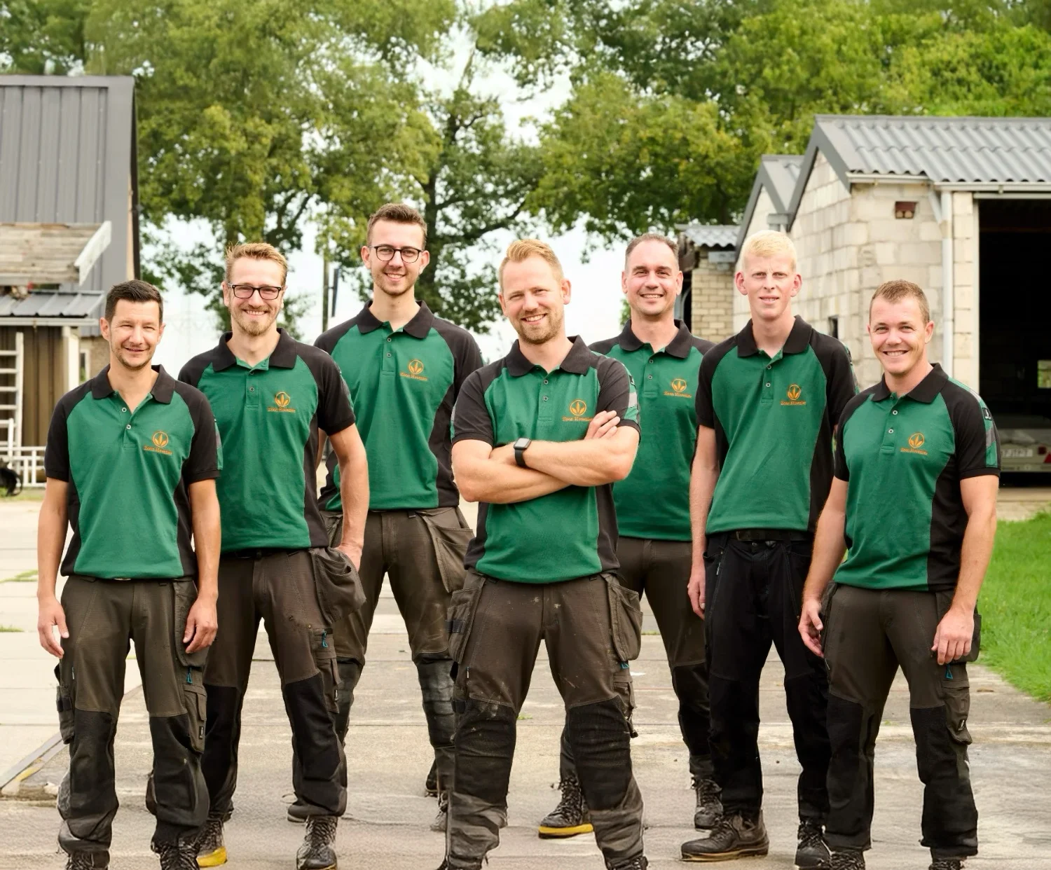 Zeven medewerkers in groen-zwarte bedrijfspolo van Zoon Hovenier staan naast elkaar op het bedrijfsterrein voor de loods